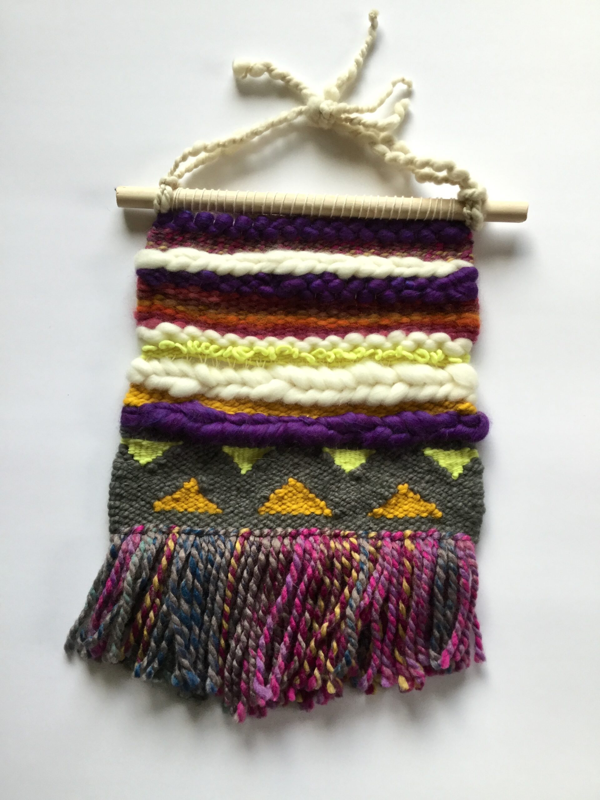 A large square weaving with stripes in autumn colors, as well as white, purple, and gray. There is a section of triangles near the bottom, and the fringe is in variegated colors. The cord is hand-spun yarn.