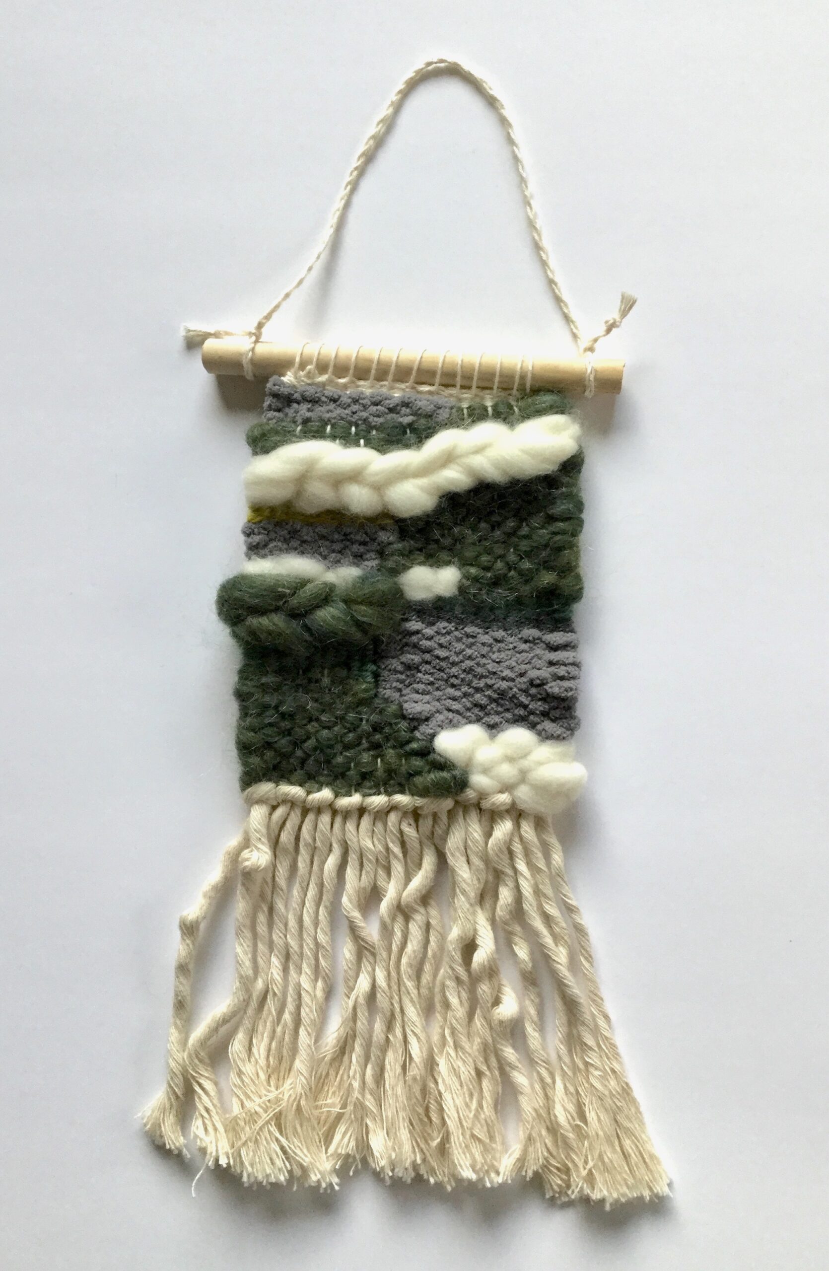 A small weaving in moss and stone colors, with a white braid near the top. It has macrame cord fringe.