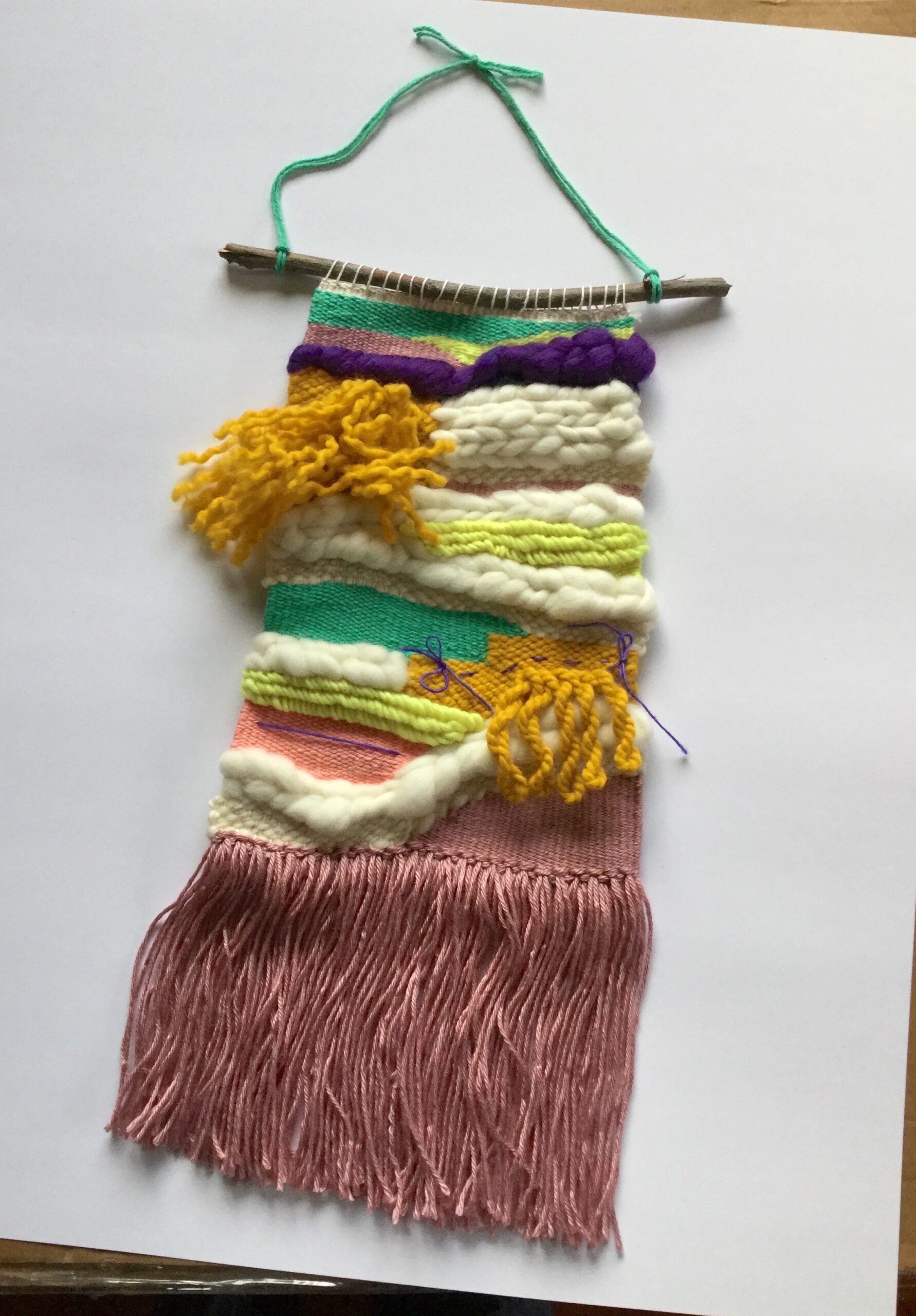 A large weaving with many textures, hung on a walnut branch. It has two shades of pink, as well as purple, teal, mustard yellow, and neon yellow. Some parts are fluffy, some parts are braided, and some are flat.