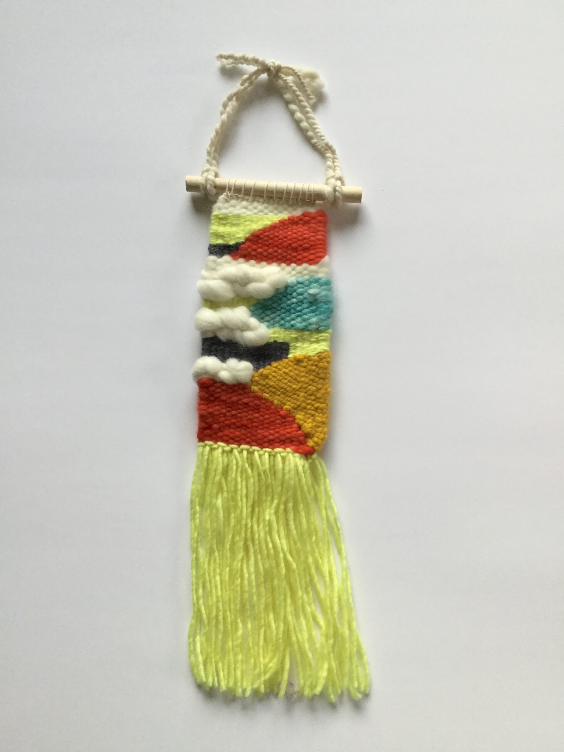 A long, thin weaving in bright primary colors (plus black and white). Many of the abstract shapes are triangles. It has yellow fringe and hand-spun yarn for the cord.