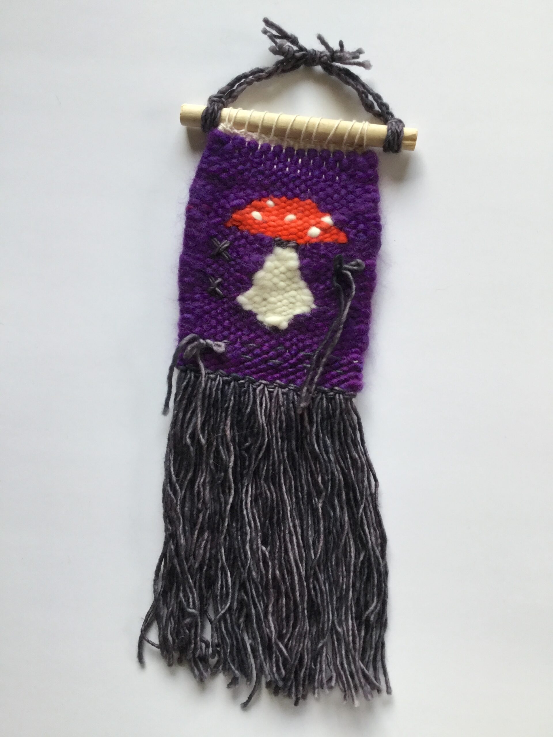 A medium sized weaving depicting an amanita muscaria mushroom on a purple background. The fringe is dark gray, with a few bows and X's in the same color scattered on the weaving.