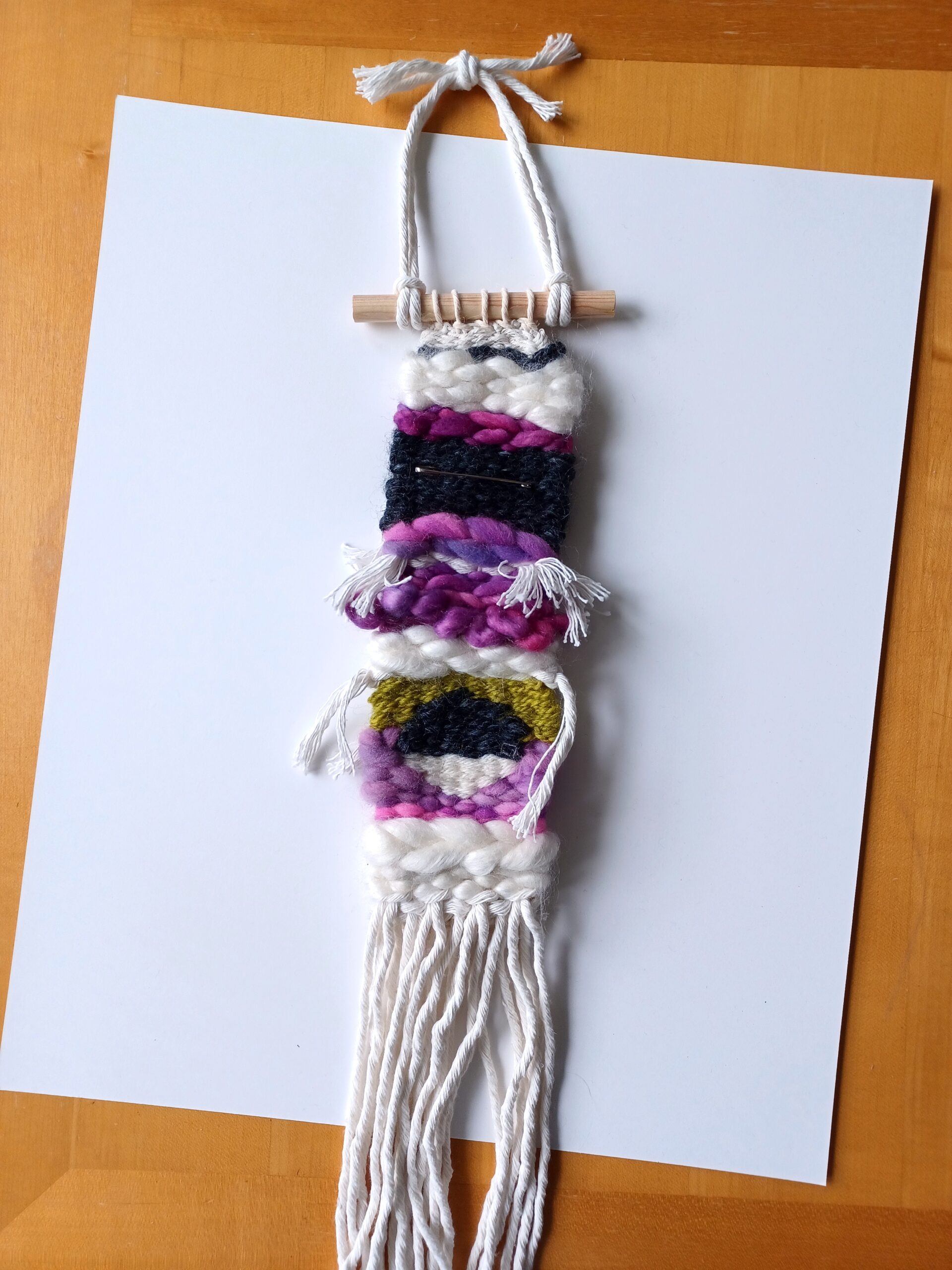 A long, thin weaving with thick cotton macrame cord fringe. It has stripes of purple, black, white, and green, with a circle divided into black and white halves near the bottom.