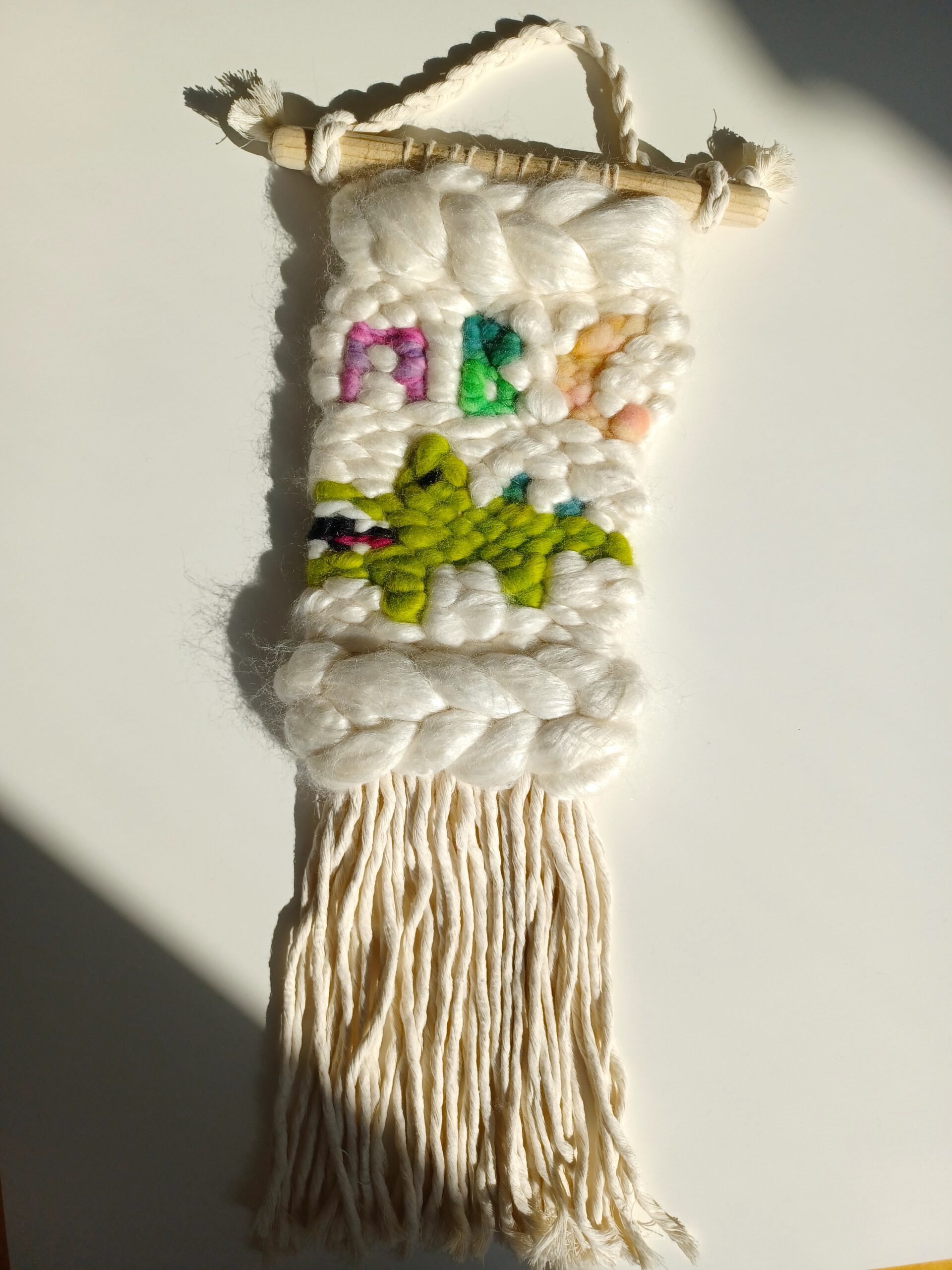 A medium-large weaving, done mostly in a shiny white wool/silk blend. There is a crocodile woven into it, with the letters "ABC" above it in the colors pink, green, and peach. The fringe is cotton macrame cord.