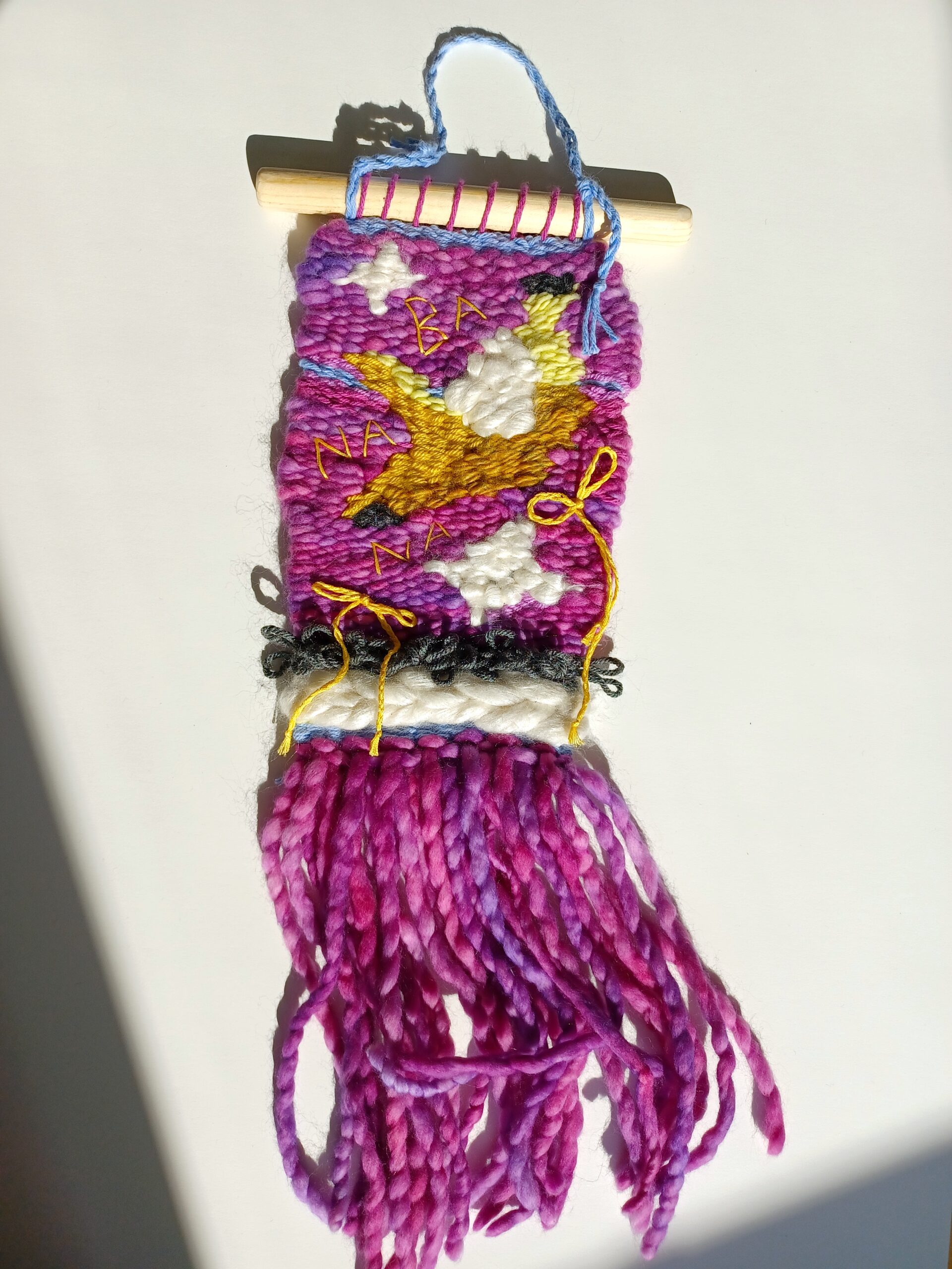 A medium weaving, with a variegated magenta background. The fringe is the same color. The main feature is a half-peeled banana design, with a couple sparkles and bows added around it, and the word "banana" embroidered on.