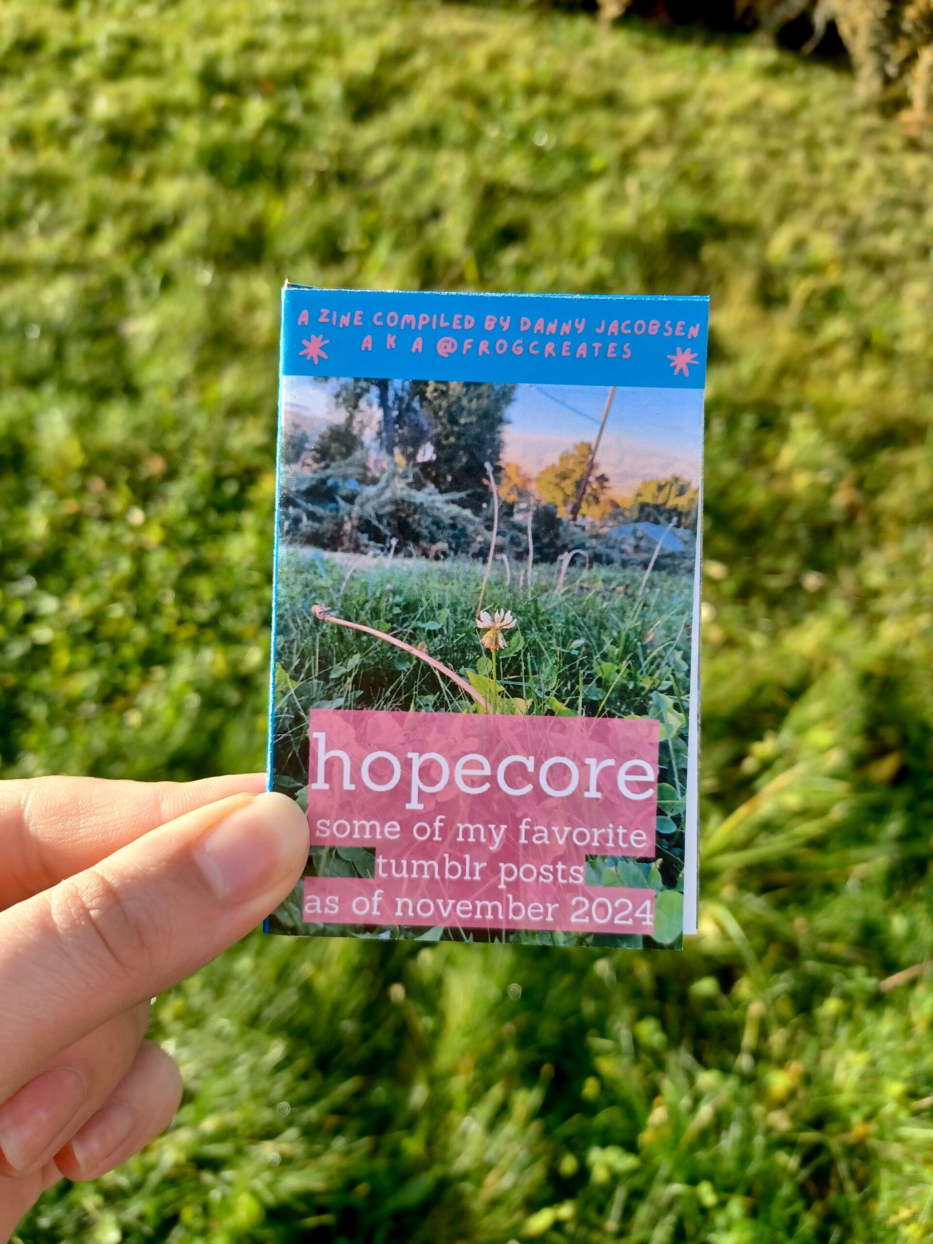 My hand holding a minizine with the title "hopecore: some of my favorite tumblr posts as of november 2024" and the subtitle "a zine compiled by danny jacobsen aka @frogcreates", in front of a grassy background.