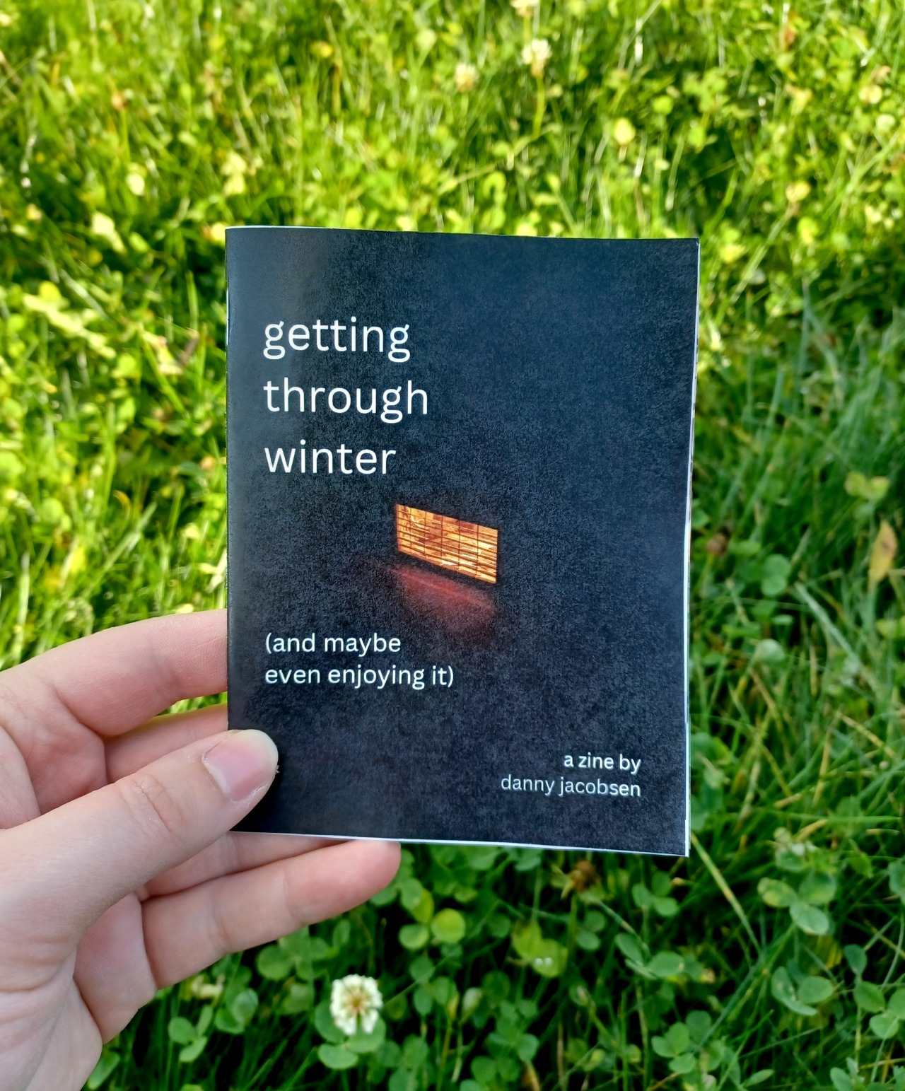 My hand holding a quarter page zine with the title "getting through winter (and maybe even enjoying it" and the subtitle "a zine by danny jacobsen", in front of a grassy background.