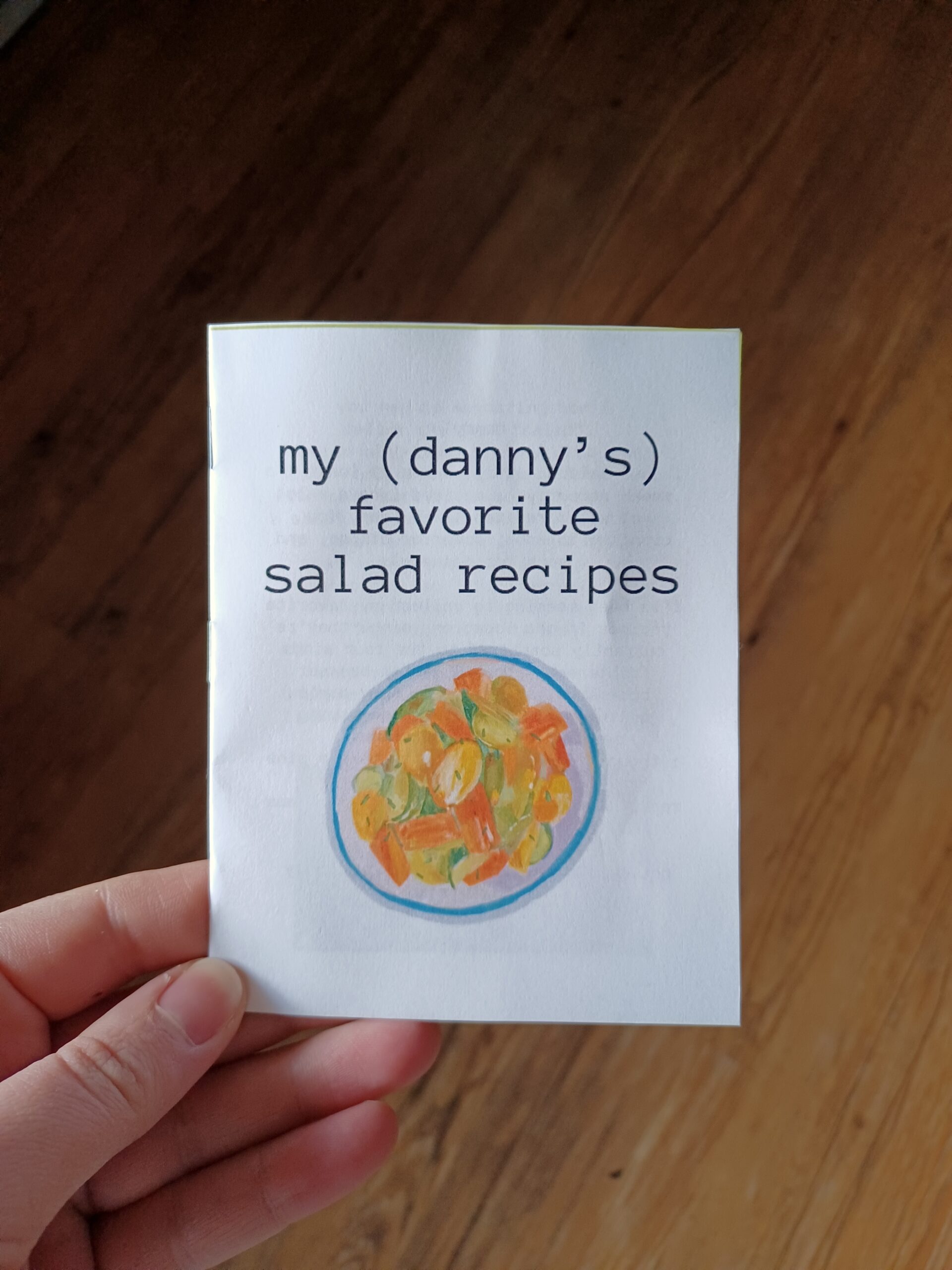 my hand holding a quarter page zine with the title "my (danny’s) favorite salad recipes" and a painting of a salad.
