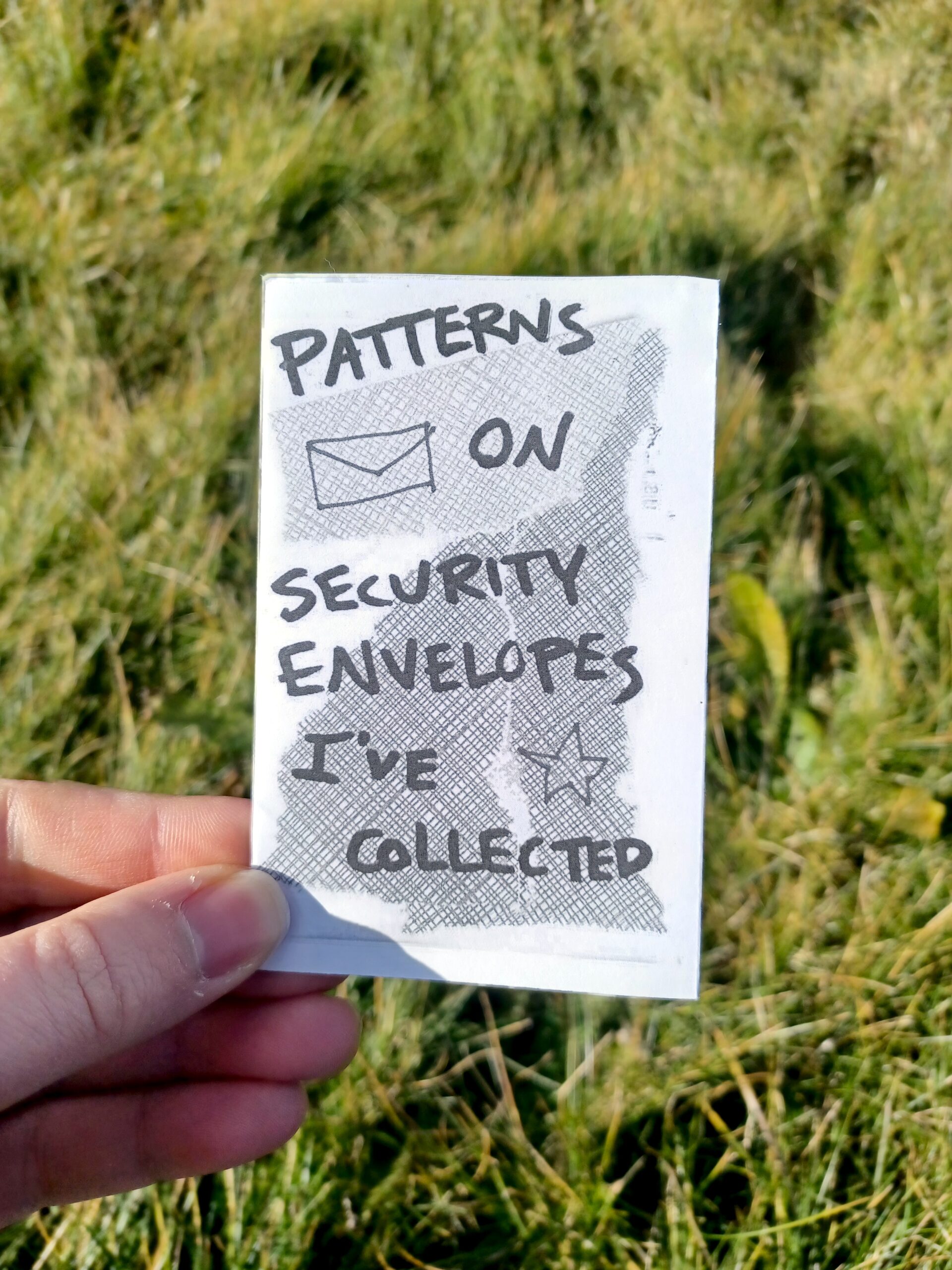 My hand holding a minizine with the title "Patterns on Security Envelopes I've Collected" in front of a grassy background.