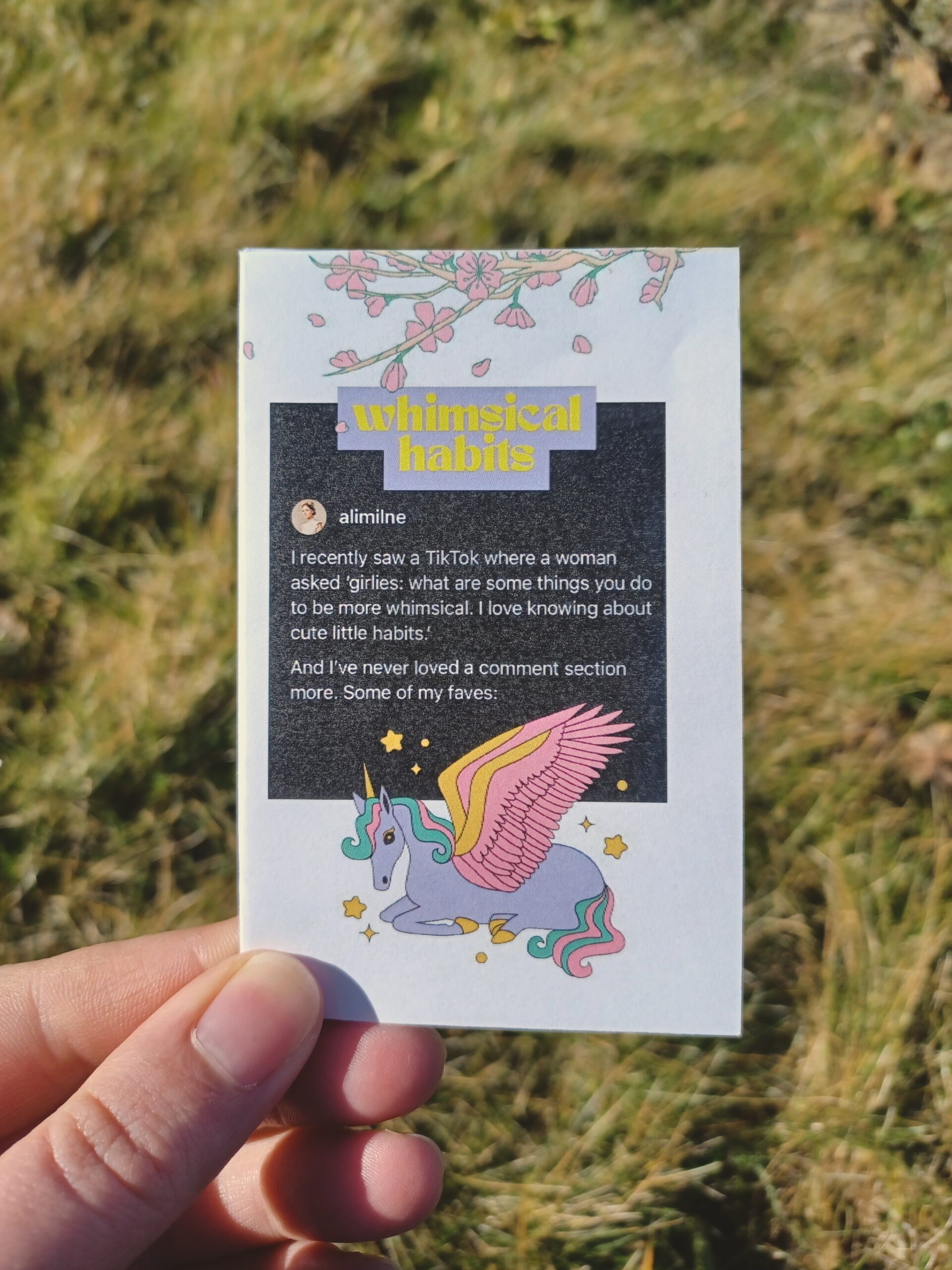 My hand holding a minizine with the title "whimsical habits" in front of a grassy background.