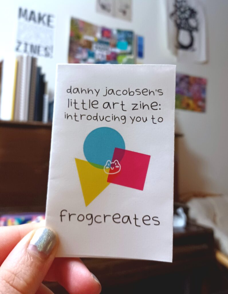 My hand holding a minizine with the title "danny jacobsen's little art zine: introducing you to frogcreates," in front of a blurry background with several posters on the wall and a piano.