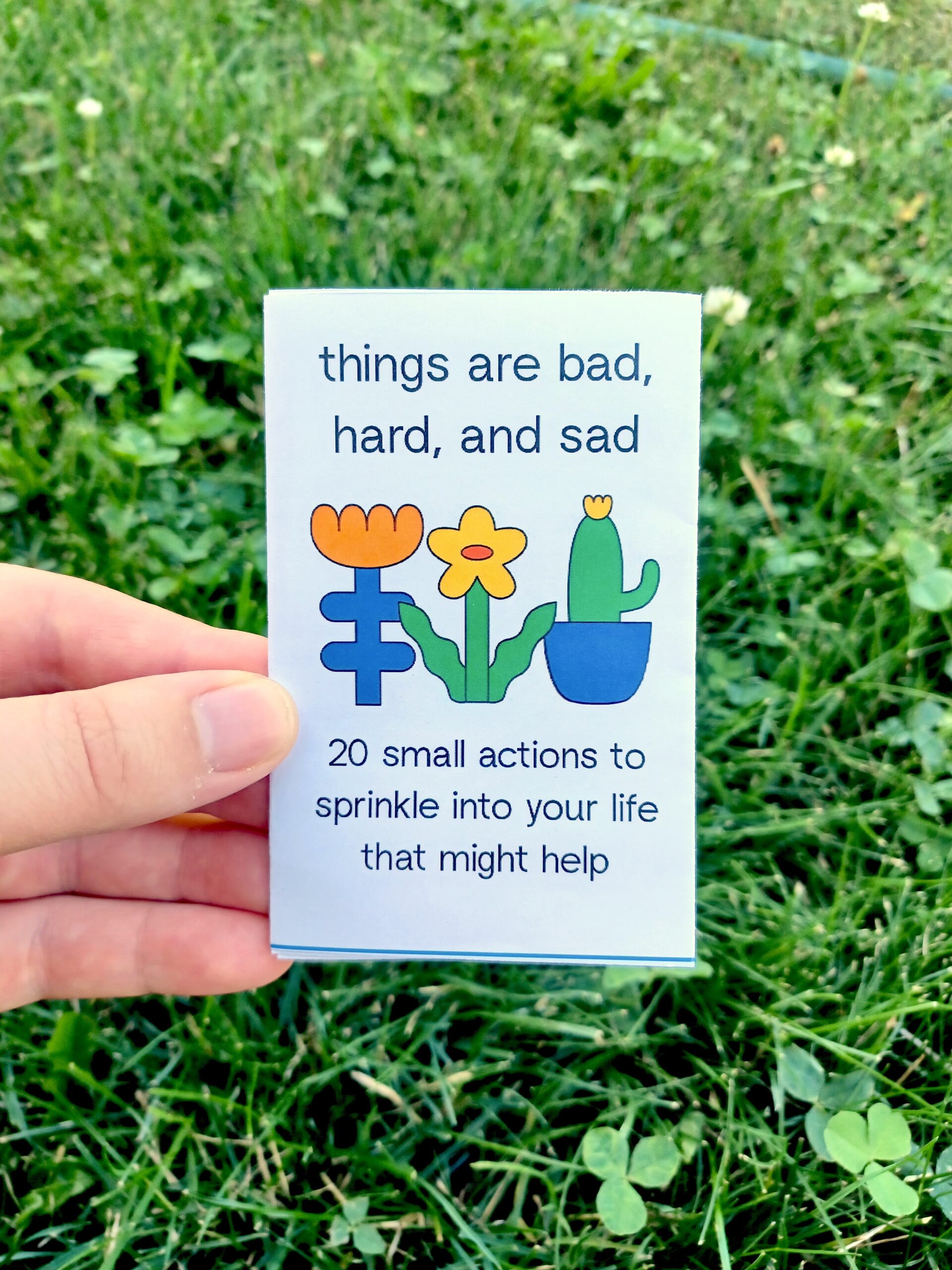 My hand holding a minizine with the title "things are bad, hard, and sad: 20 small actions to sprinkle into your life that might help" in front of a grassy background.