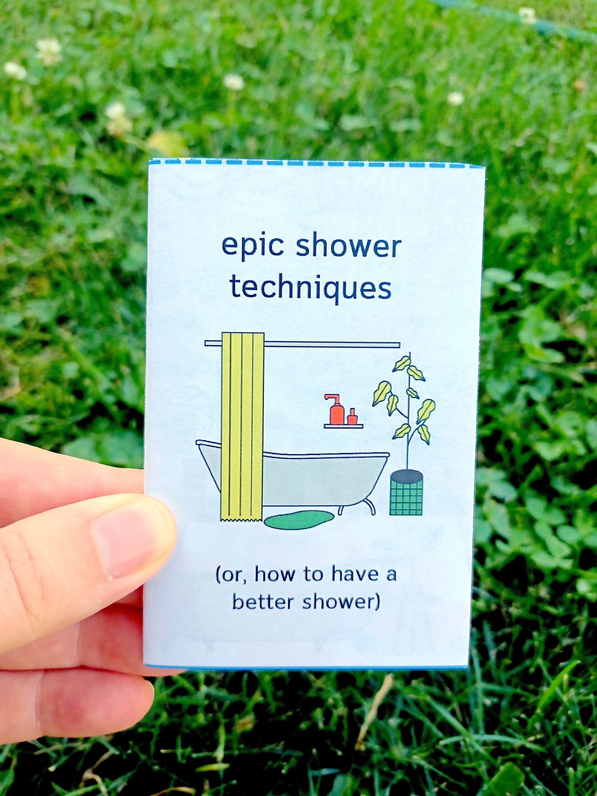 My hand holding a minizine with the title "epic shower techniques (or, how to have a better shower)" in front of a grassy background.