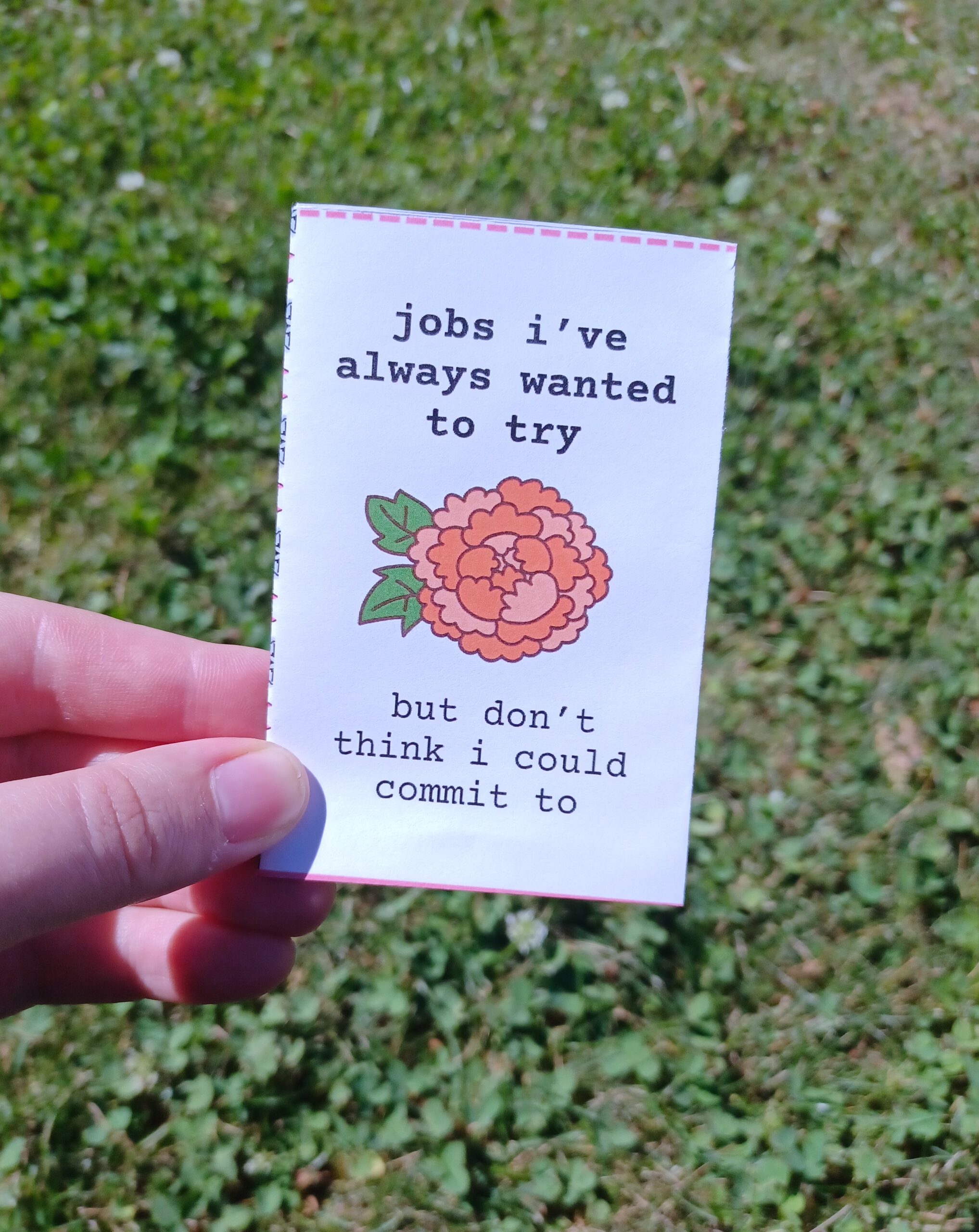My hand holding a minizine with the title "jobs i've always wanted to try but don't think i could commit to" in front of a grassy background.