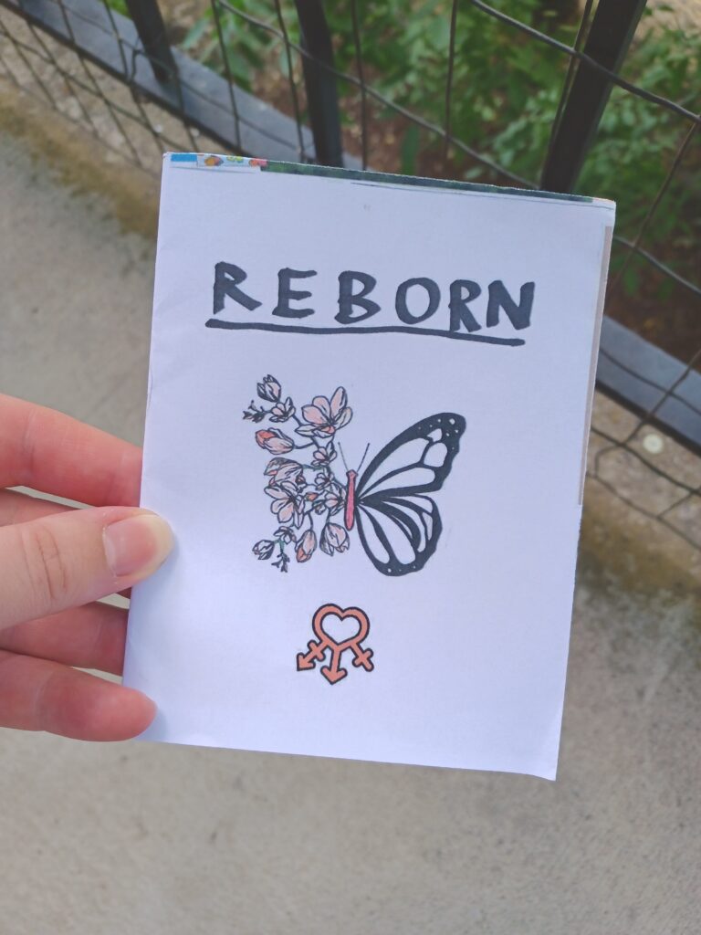 My hand holding an oversize minizine with the title "reborn" in front of a concrete background.