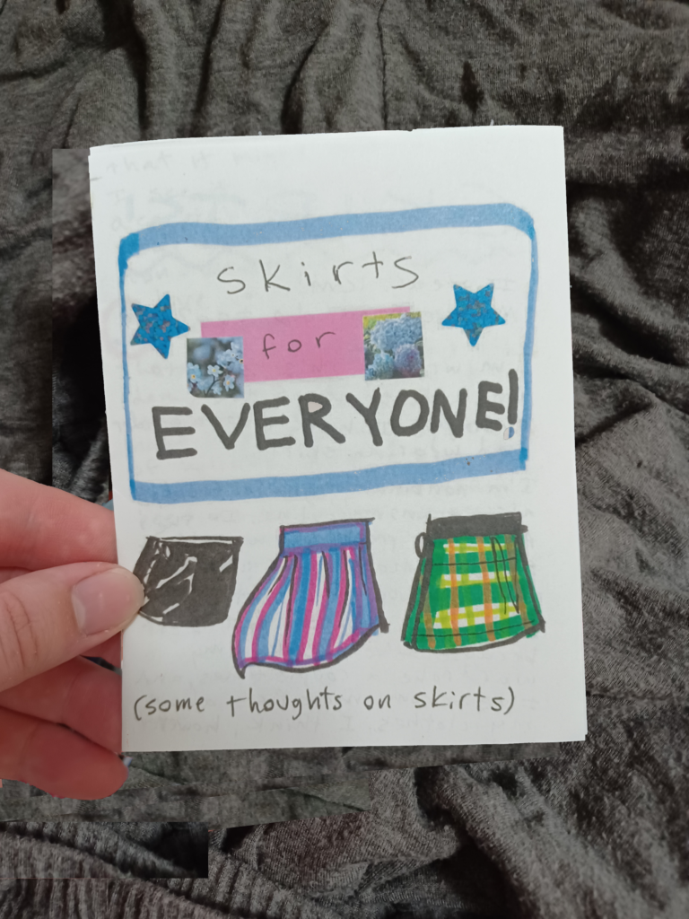 My hand holding a quarter page zine with the title "skirts for everyone!" in front of a gray fabric background.